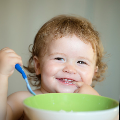 Portrait of funny little smiling boy with blonde curly hair and round cheecks eating from green plate holding spoon closeup, square picture